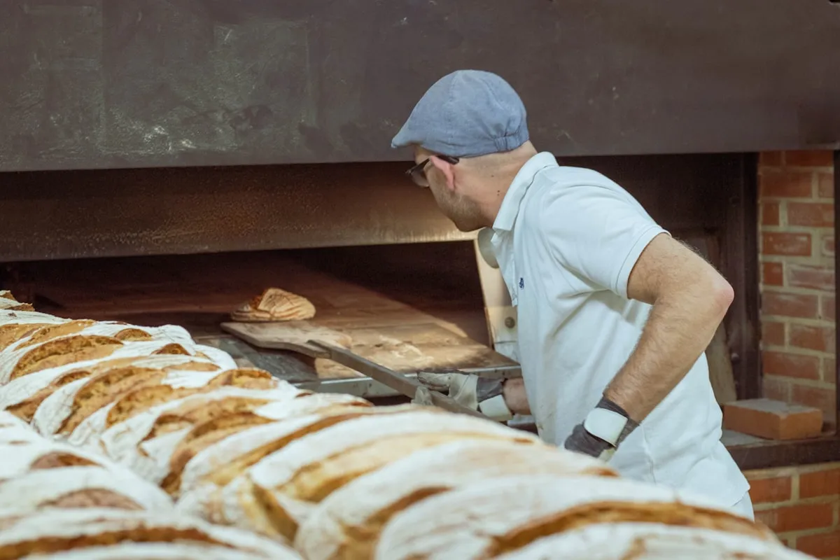 A baker in a flat cap sliding a scored loaf into a brick-fronted deck oven on a long wooden peel, with rows of floured sourdough loaves waiting in the foreground