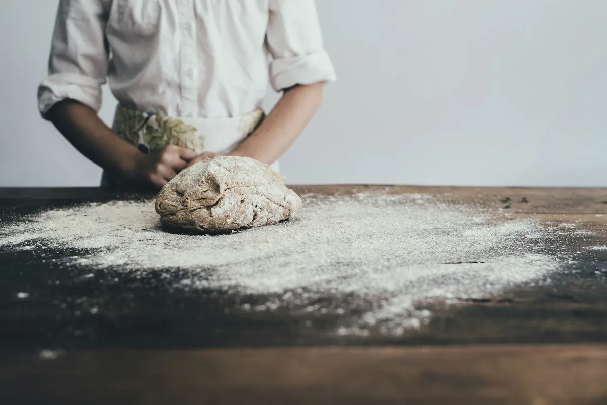 A home baker in a white shirt and a green-floral apron stands at a dark wooden table behind a freshly-floured round of dough, with flour dusted across the surface in soft, neutral natural light