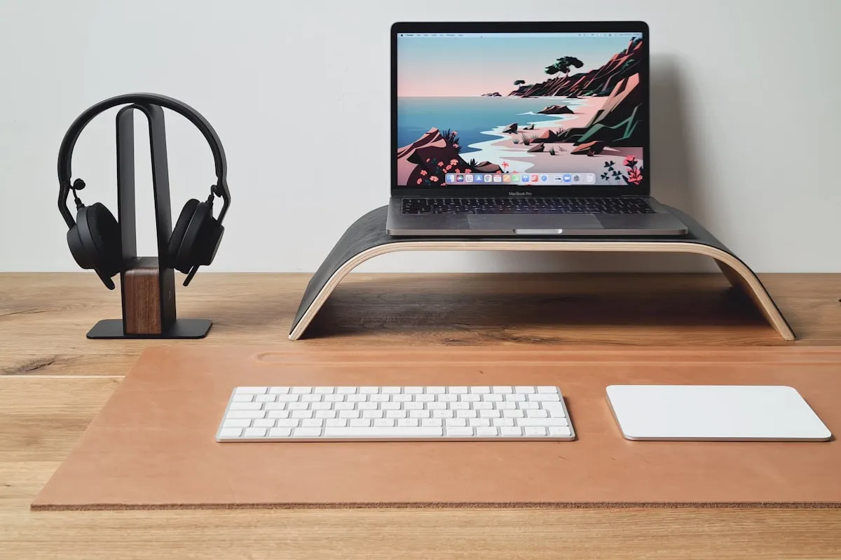 A clean modern home-office workspace with a laptop on a wooden stand, headphones on a stand, a keyboard and trackpad on a leather desk mat, and a wooden desk in soft daylight