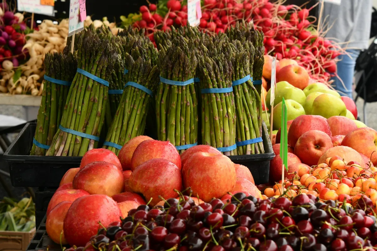 A close-up of a bright outdoor farmers market produce stand with rubber-banded bundles of fresh asparagus, stacked red and yellow apples, blushed nectarines, dark sweet cherries, golden cherries, and bunches of red radishes under handwritten paper price tags