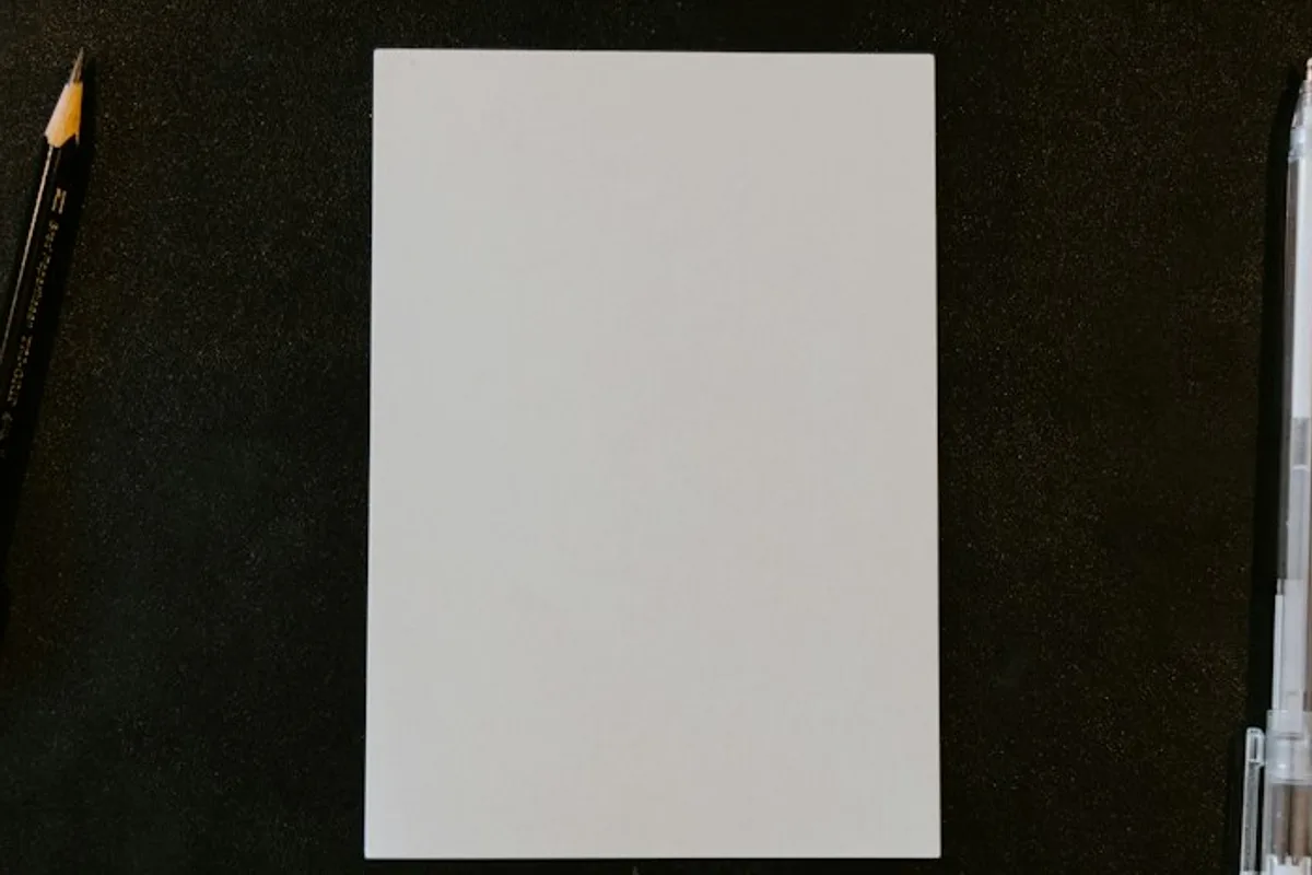 A single blank sheet of paper centered on a dark desk, shot from directly above, with a sharpened pencil resting along one edge and a pen along the other