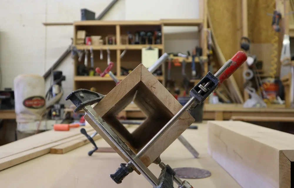 A wooden workpiece held square in a corner clamp and a bar clamp during glue-up, on a bench in a working woodworking shop