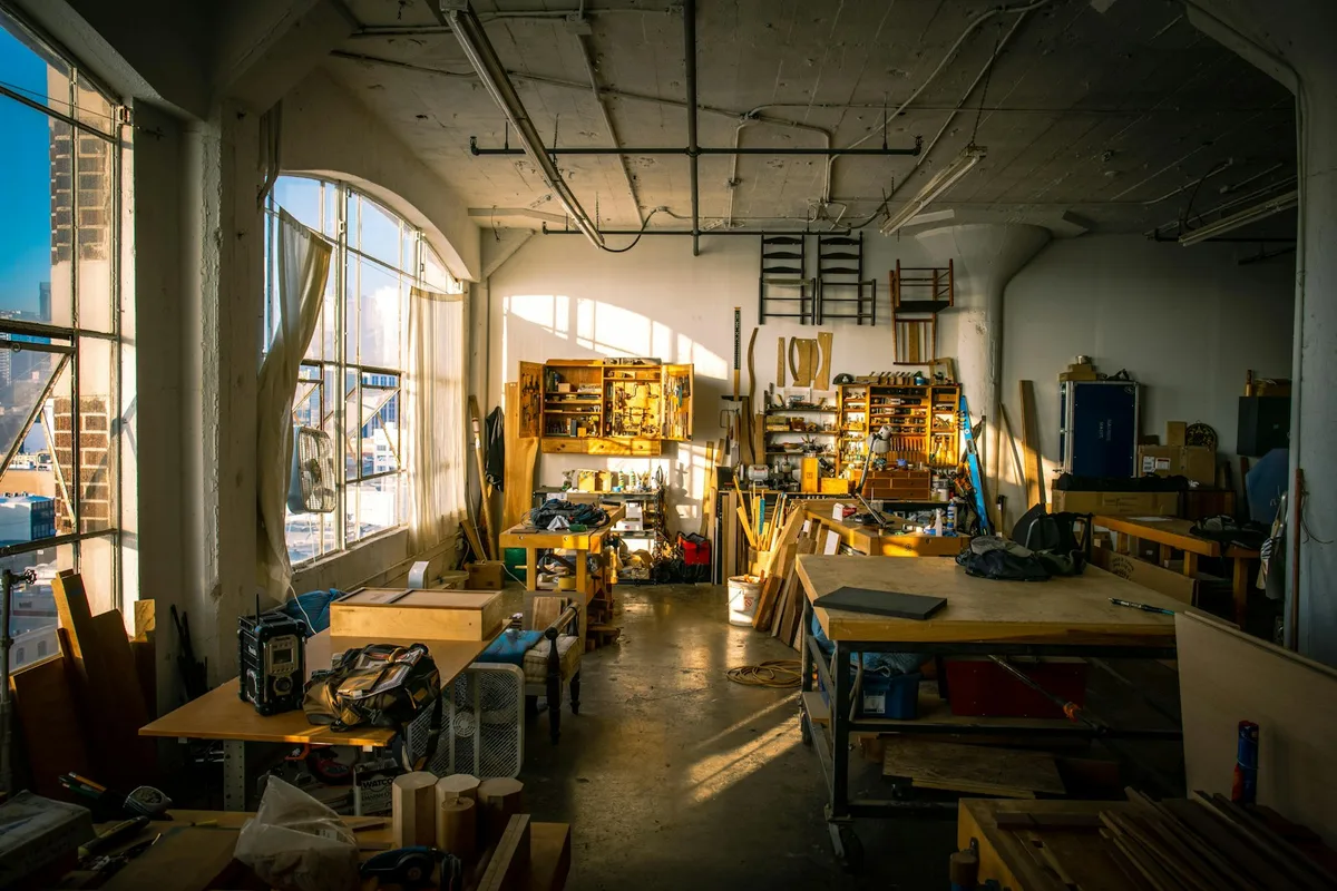 A one-person woodworking studio in golden afternoon light — long workbenches, stacked lumber, wall-mounted tool storage, and tall arched windows looking out onto a city skyline
