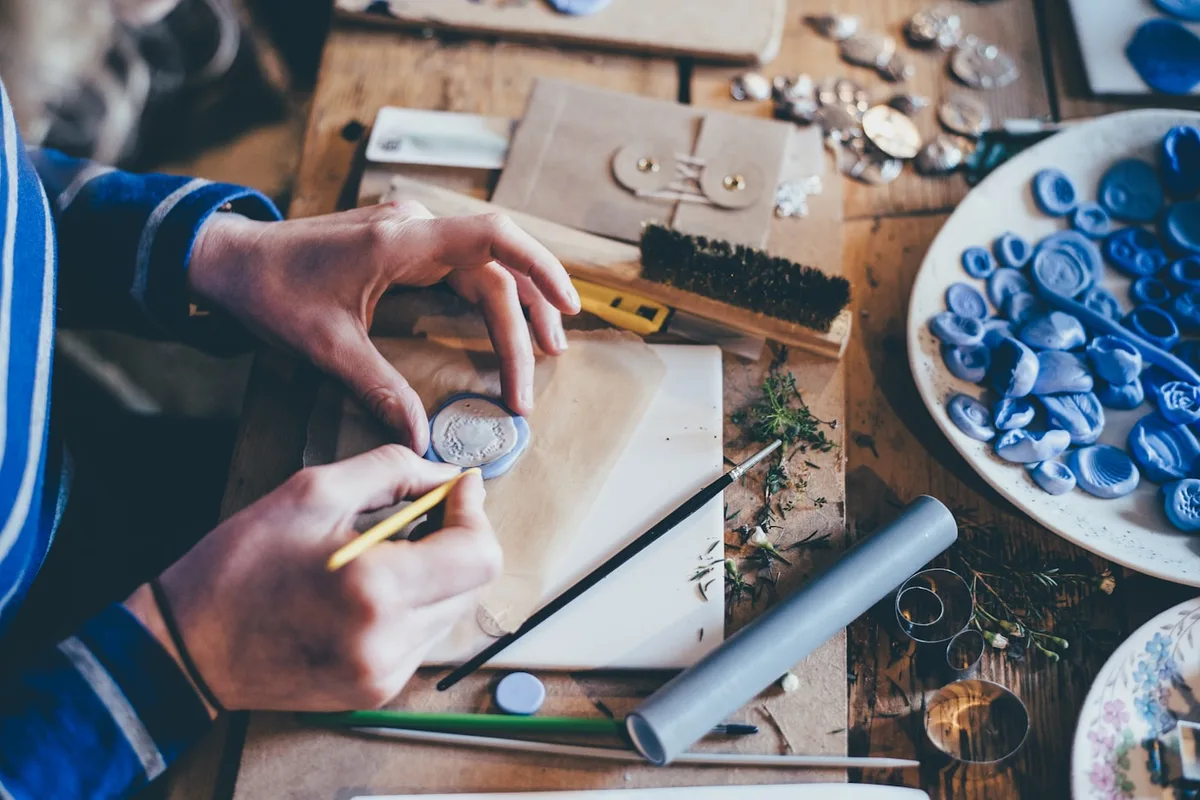 Hands at a wooden workbench shaping a small clay piece with a fine tool, surrounded by molds, brushes and metal findings