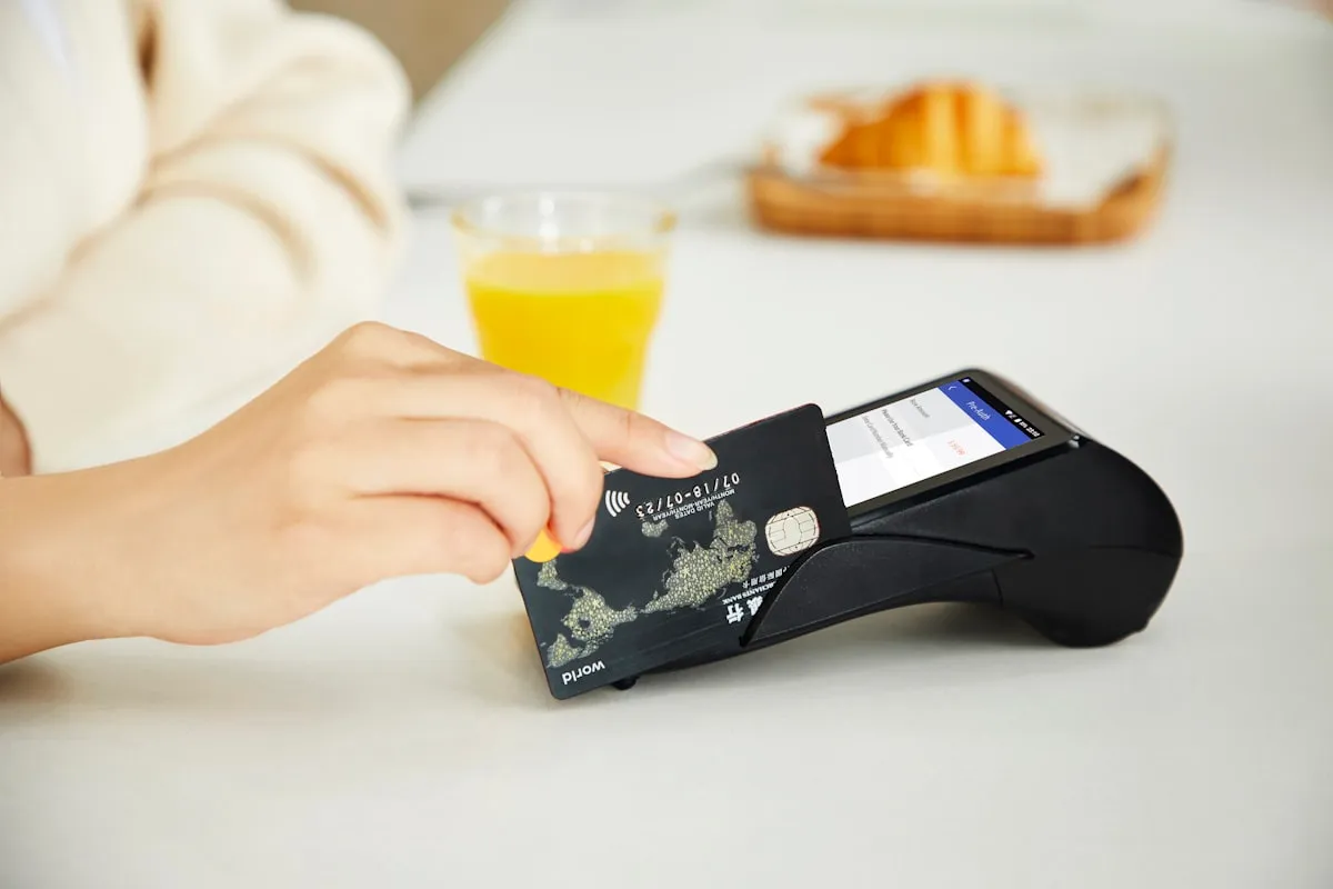 A hand inserting a dark chip card into a black countertop card terminal on a pale table, with a glass of juice and a croissant blurred behind