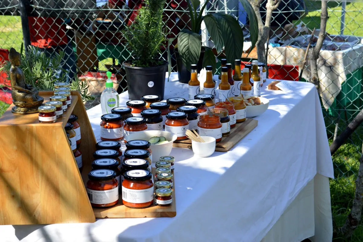 A farmers market vendor table arranged with rows of small dark-capped preserve jars on a wooden stepped display, larger jars of orange tomato sauces and chutneys, a line of dark-glass hot sauce bottles, and potted rosemary on a white linen tablecloth in an outdoor market setting under afternoon light