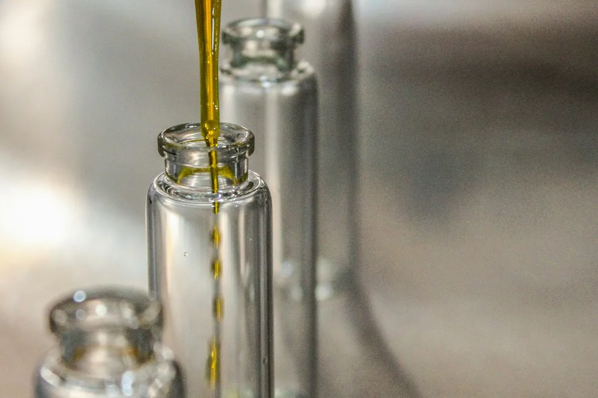 Amber aromatic material dripping from a pipette into a small glass vial on a perfumer's workbench