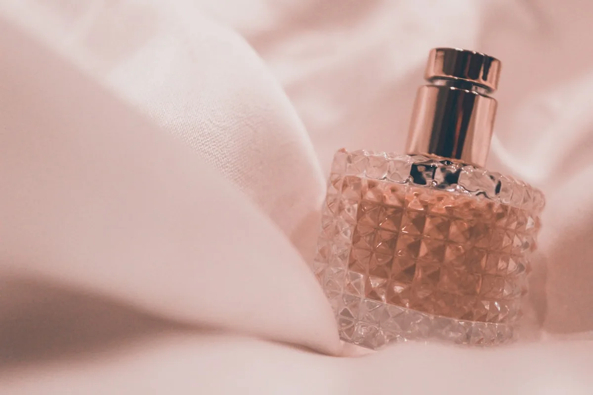 A faceted glass perfume bottle with a rose-gold atomizer resting on soft blush-pink fabric