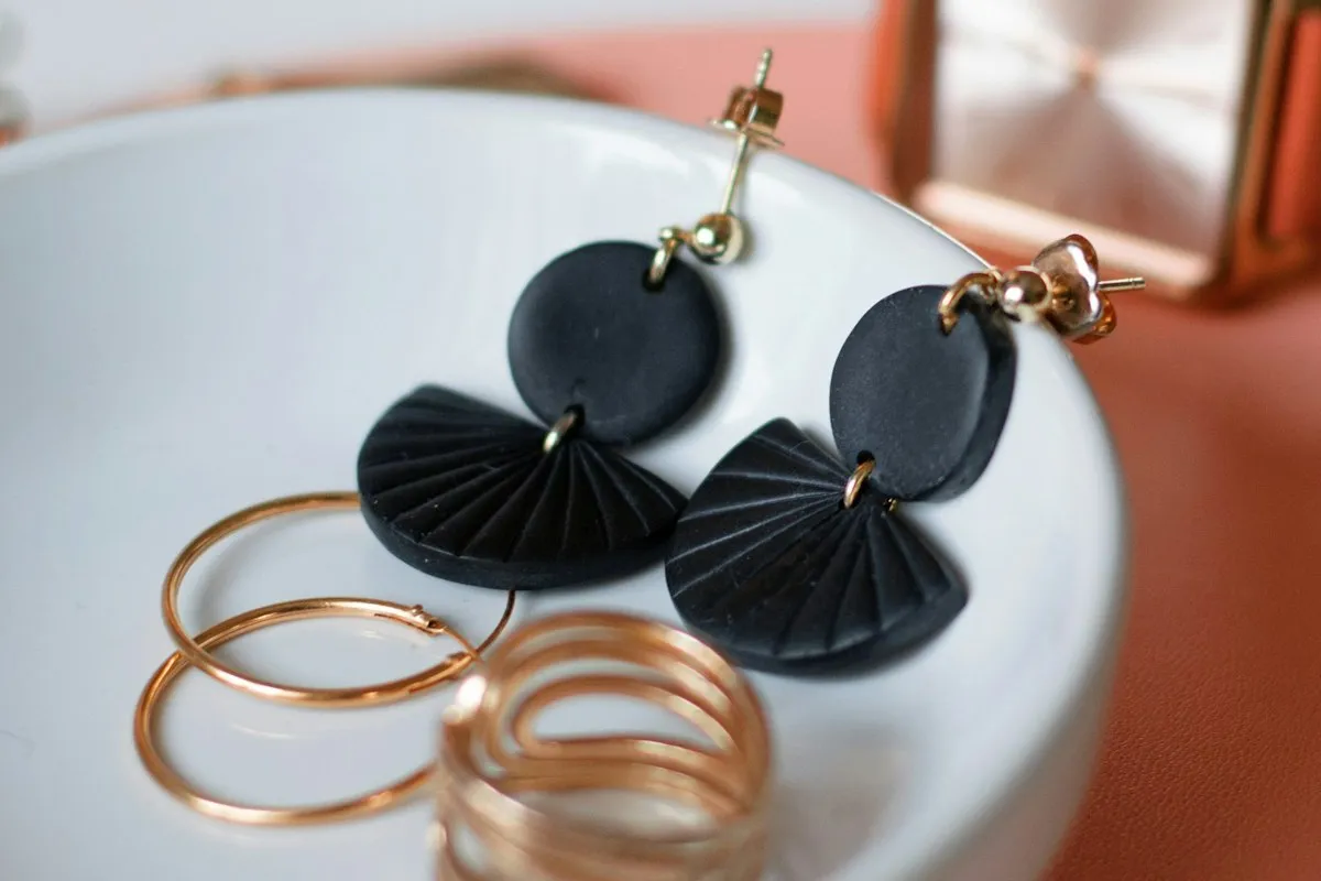A pair of black polymer clay fan-shaped statement dangle earrings — the style analyzed in Worked Example 2 — resting in a white ceramic dish beside gold rings