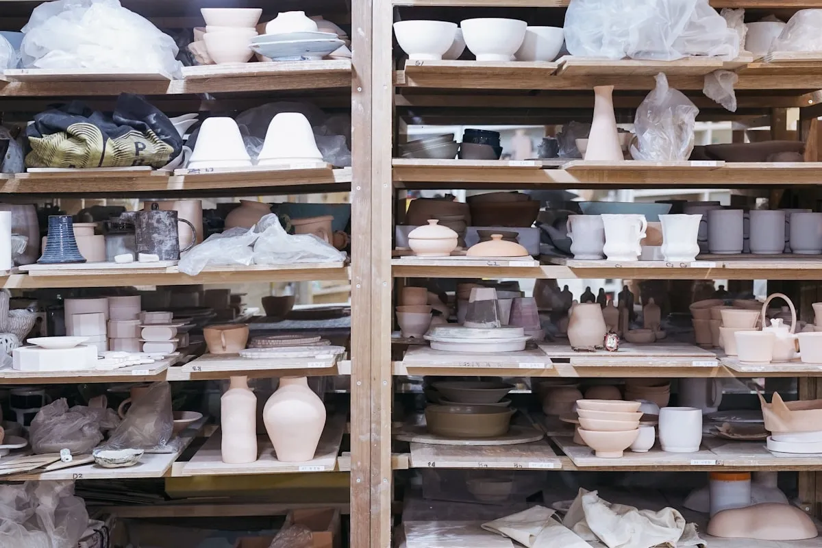 Wooden studio shelves filled with rows of unfired bisqueware — bowls, vases, cups, lidded jars, and plates — drying and waiting for the bisque fire
