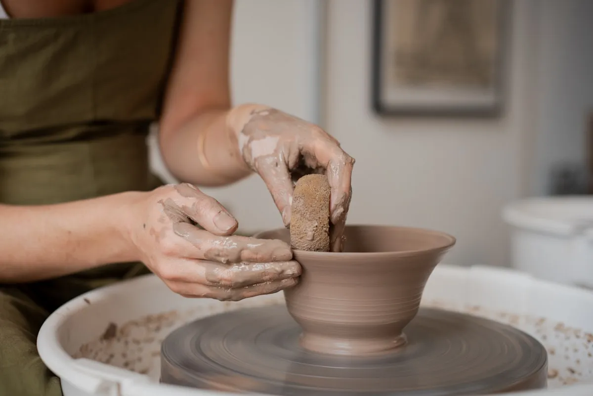 Clay-covered hands shaping a bowl on a spinning pottery wheel with a sponge, in a bright modern studio