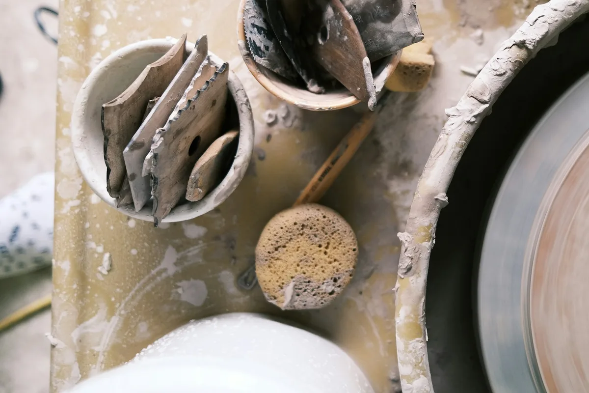 Overhead view of a potter's splash pan: wooden ribs and metal scrapers in two clay-caked cups, a wet sponge, and a plate turning on the wheel head