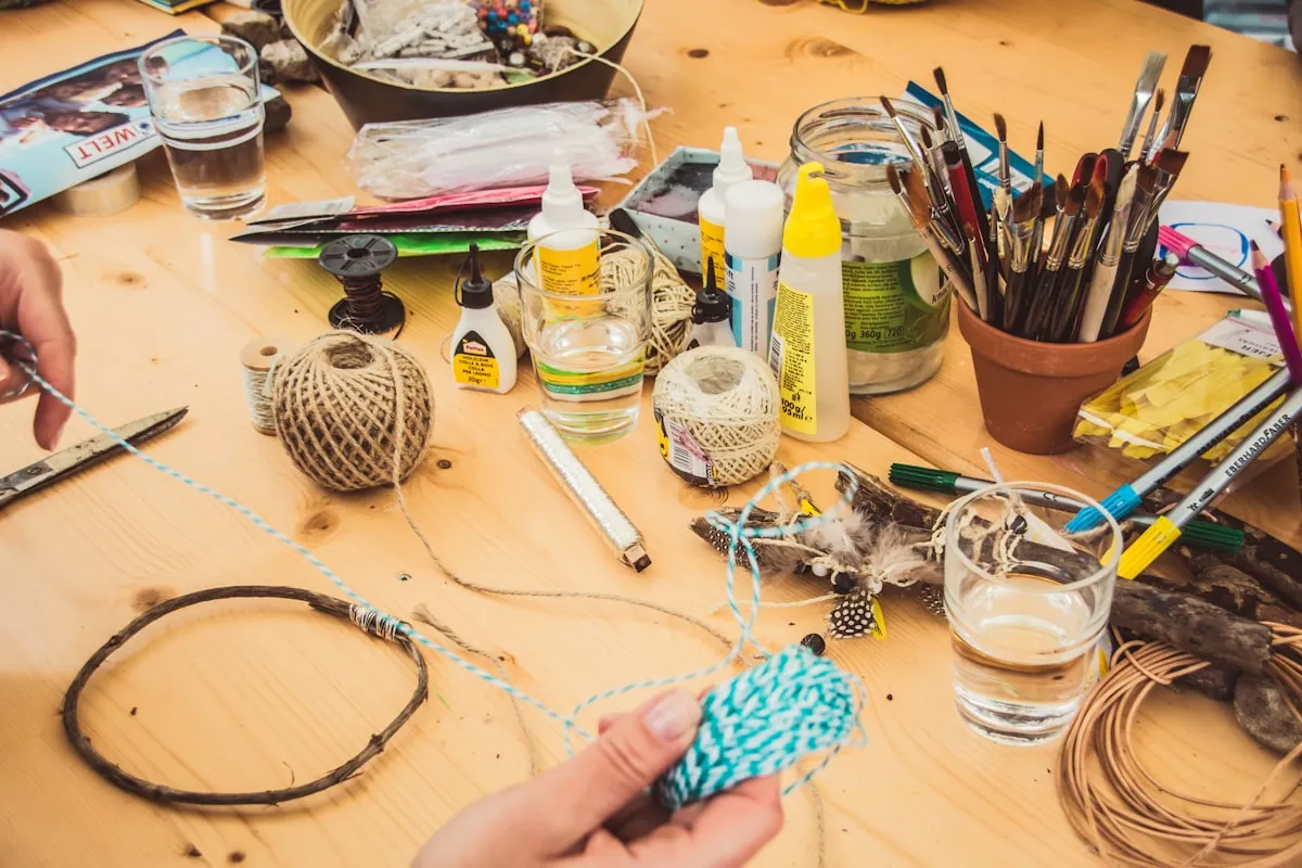 A craft workshop table covered with twine, paintbrushes, glue, feathers, and colored pencils as a pair of hands works on a project