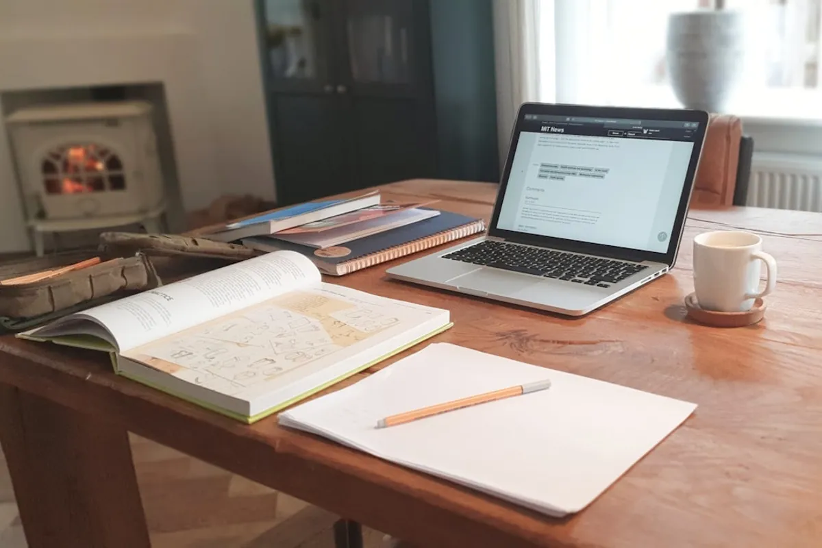 A warm wooden home-office desk with an open laptop, an open reference book, a blank notepad with a pencil, and a coffee mug beside a wood-burning stove