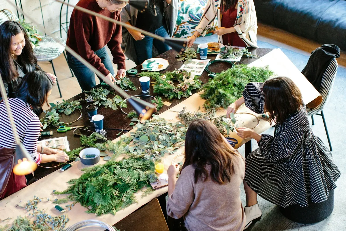 Overhead view of a group seated around a long table at a hands-on craft workshop, working with greenery and materials spread across the table