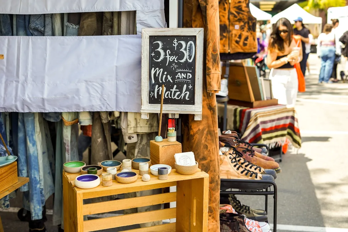 A craft fair vendor booth with a handwritten chalkboard sign reading "3 for $30 Mix and Match" propped above a wooden crate display of small handmade ceramic bowls in earth-tone glazes, with browsing shoppers and other vendor stalls blurred in the background
