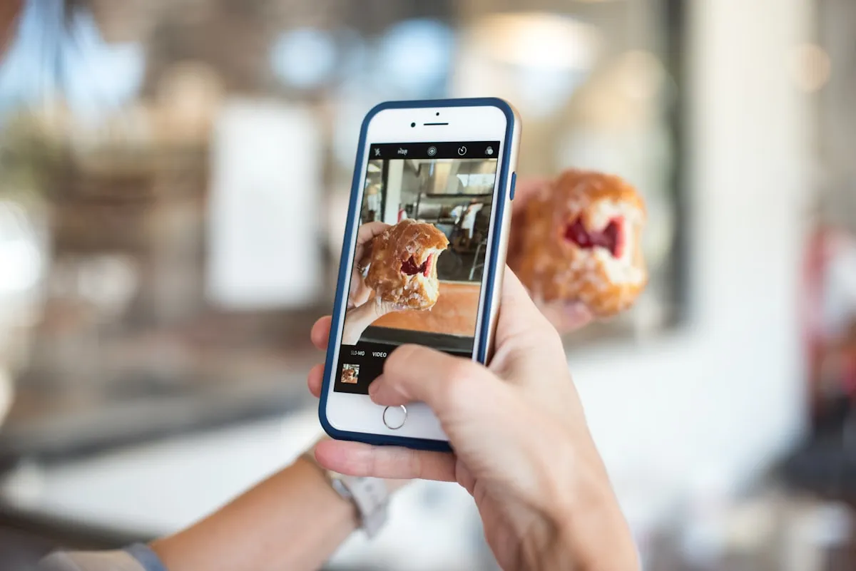 Person holding a smartphone to photograph a pastry, with the phone screen showing the camera viewfinder and a soft bokeh background