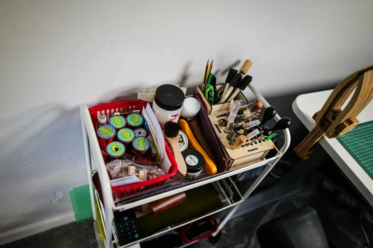 A white rolling cart in a leatherworking studio, its baskets packed with spools of waxed thread, jars of finish, stitching chisels and edge tools beside a cutting mat