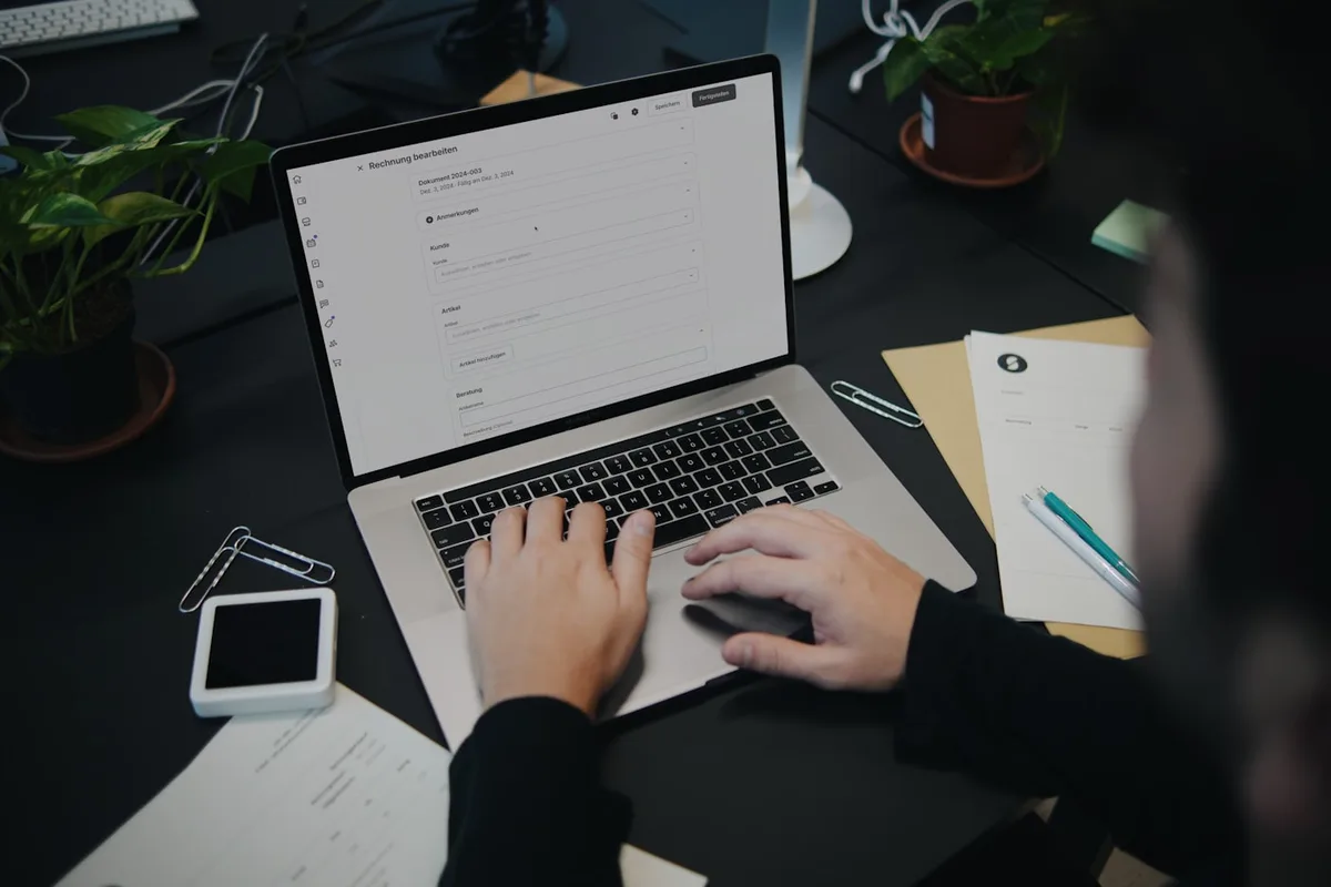 Over-the-shoulder view of a person typing on a laptop that displays an invoicing app, at a dark desk with invoice papers, a pen, paperclips, and potted plants
