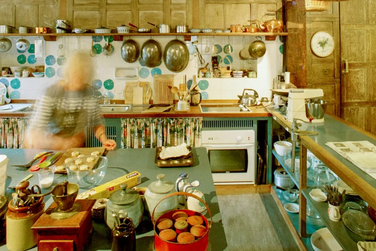 A home baking kitchen with hanging copper pans, a tray of unbaked biscuits on a blue work table, and a baker moving past in a blur