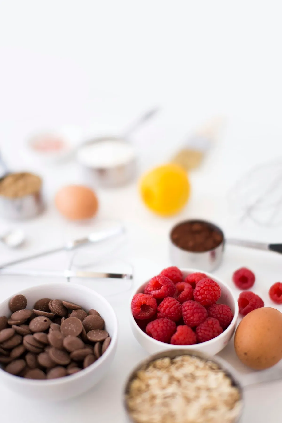 Baking ingredients including chocolate chips, raspberries, eggs, and cocoa measured into small white bowls on a white surface