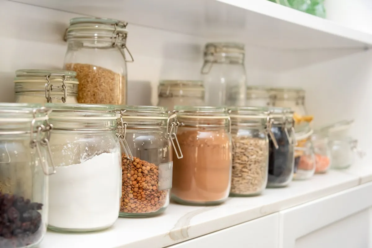 A row of clear glass clamp-lid jars filled with flour, lentils, cocoa, oats, and other baking staples lined up on a bright white pantry shelf