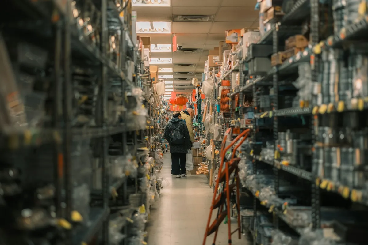 Two shoppers far down a narrow supply-store aisle lined floor to ceiling with packaged hardware and housewares on steel shelving, a wooden stepladder propped in the foreground
