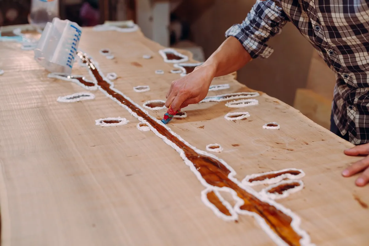 A maker's hands working amber epoxy resin and pigment into the routed channels of a pale wood slab on a workshop bench — an early step in making a river-style resin coaster