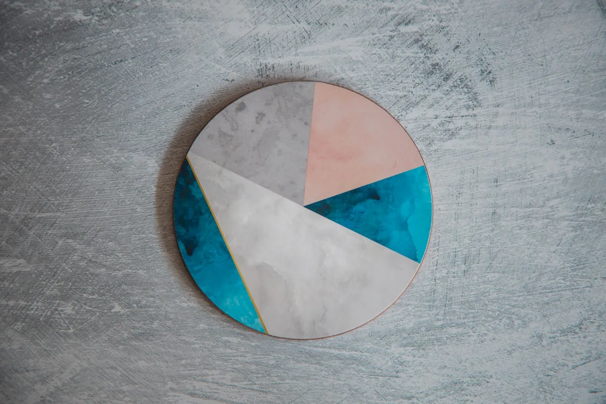 A single round resin coaster with grey, blush-pink, and teal geometric segments, resting on a textured pale-grey concrete surface