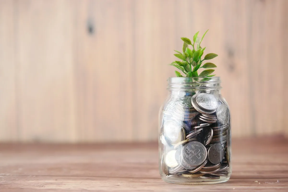 A glass jar filled with silver coins with a small green plant sprouting from the top, sitting on a wooden surface against a warm wooden background