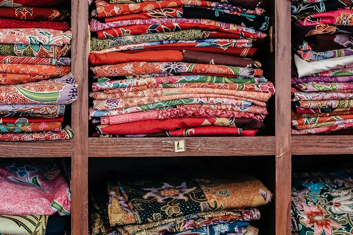 Colorful folded fabrics in reds, greens, and batik prints stacked neatly on dark wooden shelves