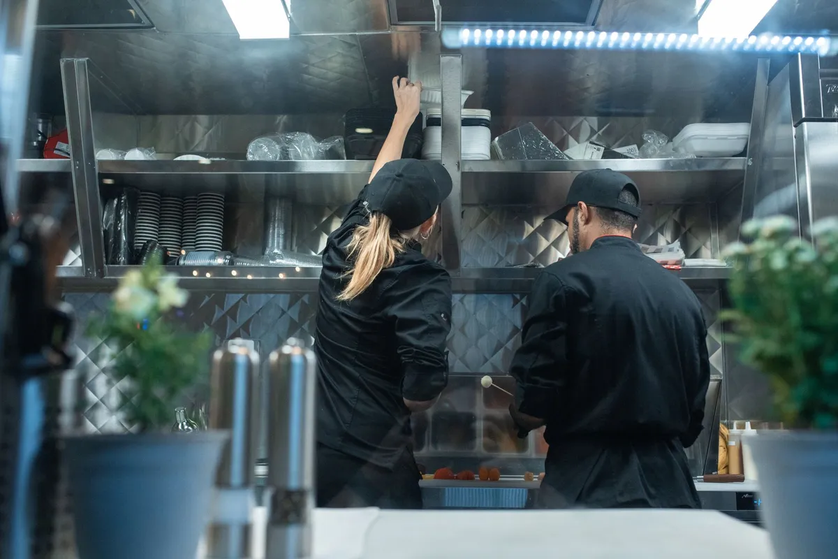 Two cooks in black jackets and caps working with their backs to the camera under stainless steel shelves stacked with containers in a commercial kitchen