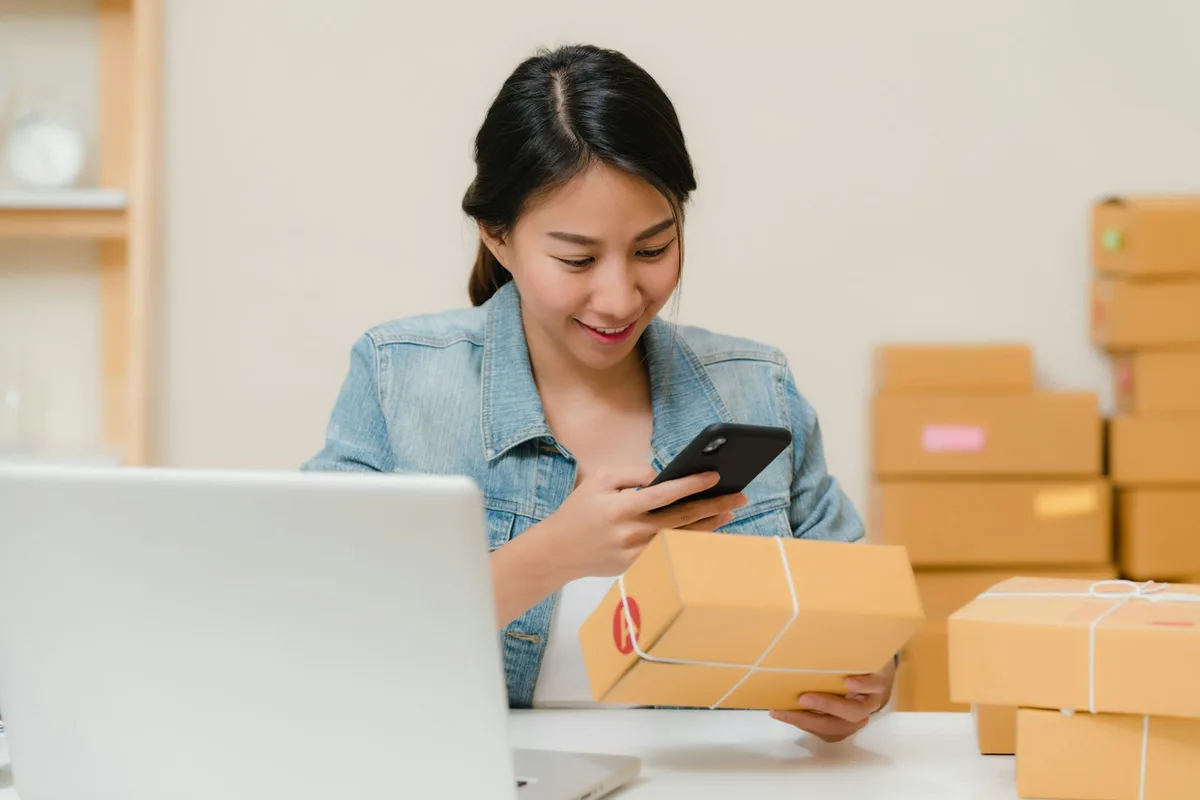 A small-business seller at a desk with an open laptop, holding a string-tied kraft parcel in one hand and a phone in the other, with stacks of packed boxes waiting alongside