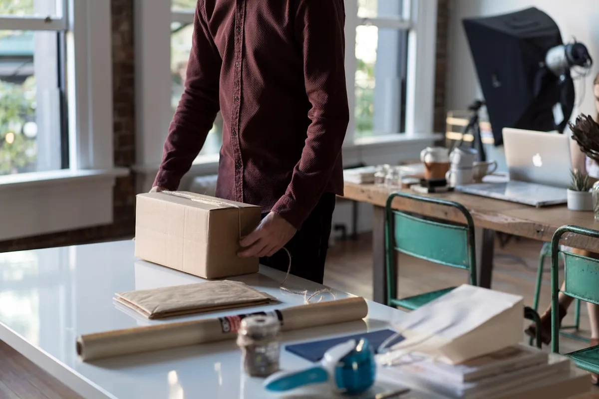 A maker standing at a bright studio table with a small corrugated box, a roll of kraft paper, twine and tape laid out ready to pack an order
