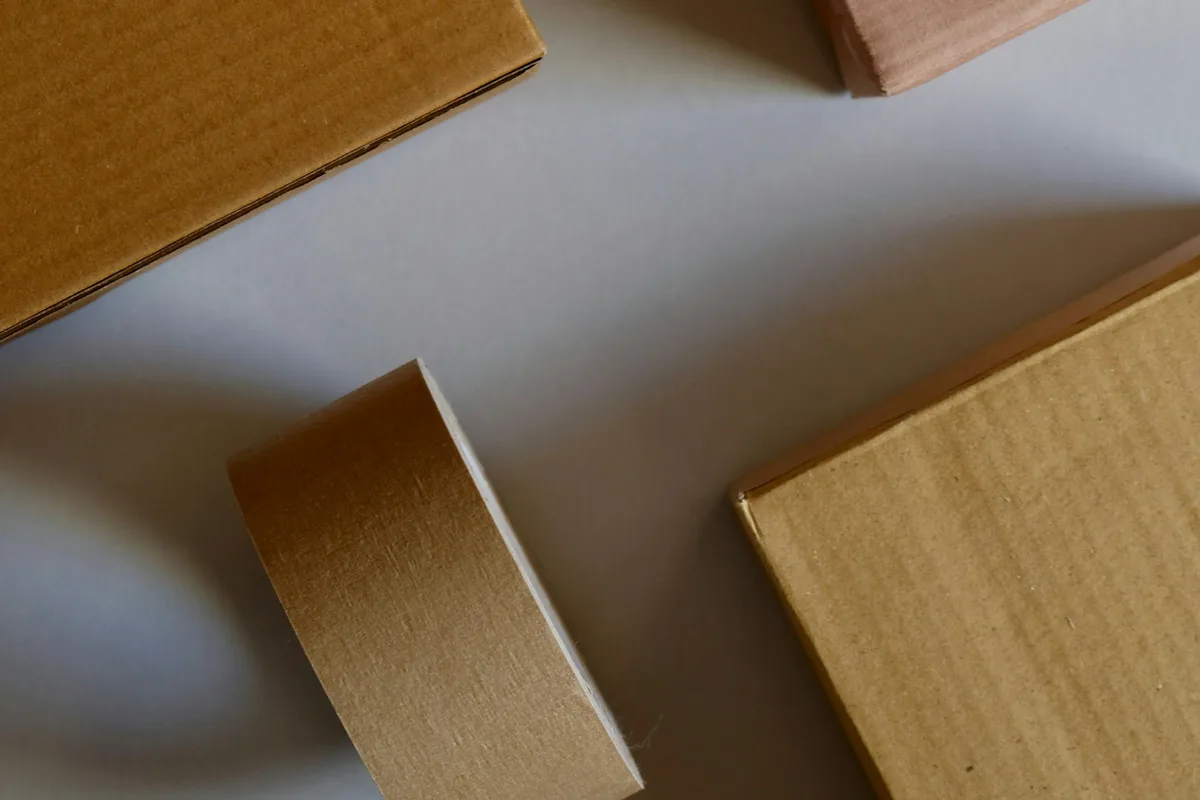An overhead flat lay of three kraft cardboard shipping boxes and a roll of paper packing tape arranged on a pale gray surface