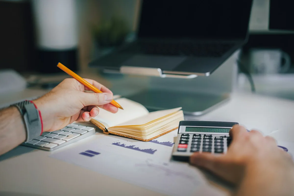 A close-up of two hands at a desk — one holding a yellow pencil over a small open notebook, the other working a black calculator, with a printed bar-chart report on the desk between them and a laptop, keyboard, and warm reading lamp softly out of focus in the background