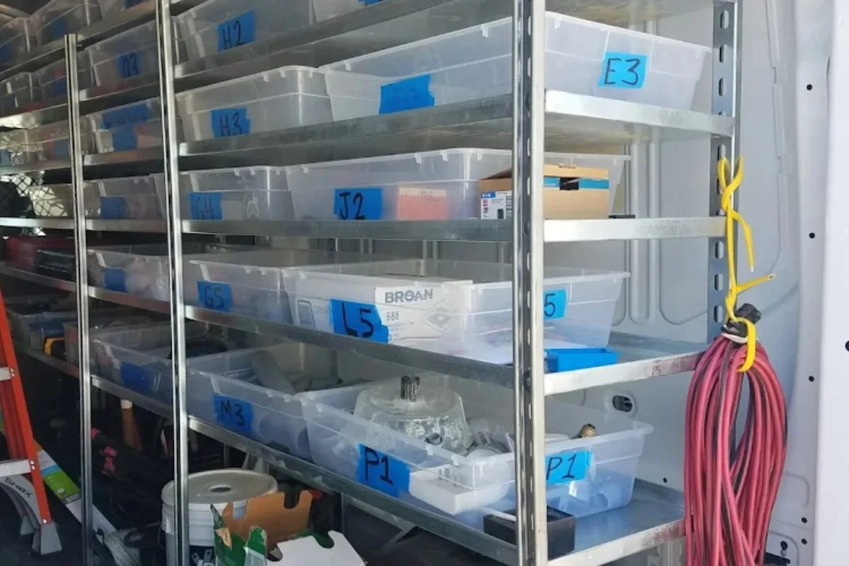Rows of clear plastic storage bins arranged on metal shelving, each labeled with a short alphanumeric code on blue painter's tape