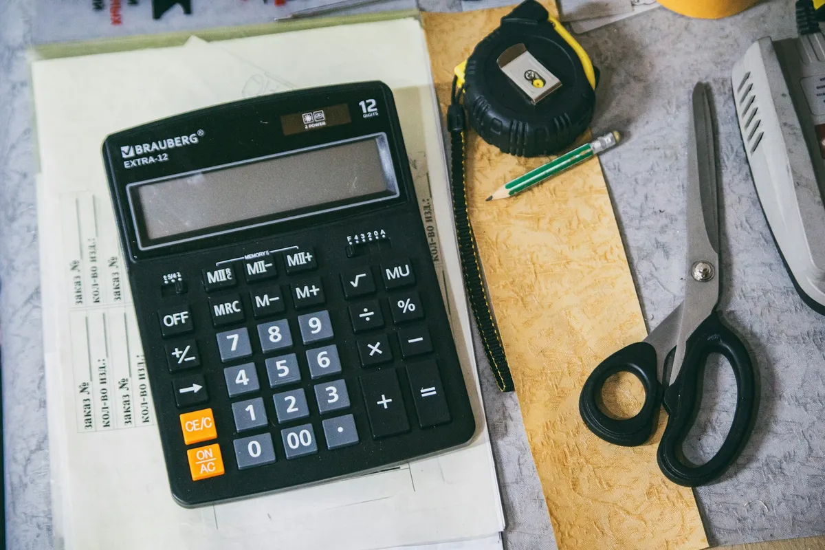 Overhead view of a calculator resting on a stack of order forms on a workbench, beside a tape measure, a pencil and a pair of fabric scissors