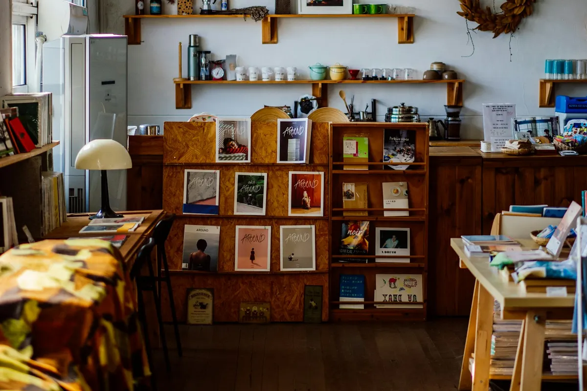A small independent maker shop interior with handmade magazines and zines arranged on wooden shelves, a vintage lamp, mugs lined up on a high shelf, and warm afternoon daylight on a wooden floor