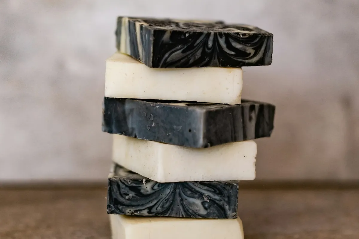 A stack of six handmade cold-process soap bars — alternating cream-white and charcoal-swirl — balanced on a weathered wooden surface against a soft neutral concrete backdrop