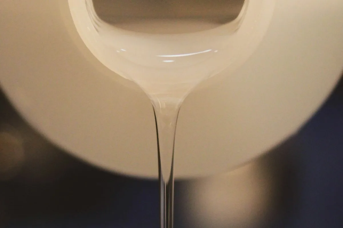 A creamy stream of liquid pouring from a wide white pot down into a metal vessel against a deep blue background — the visual moment of a cold-process soap pour