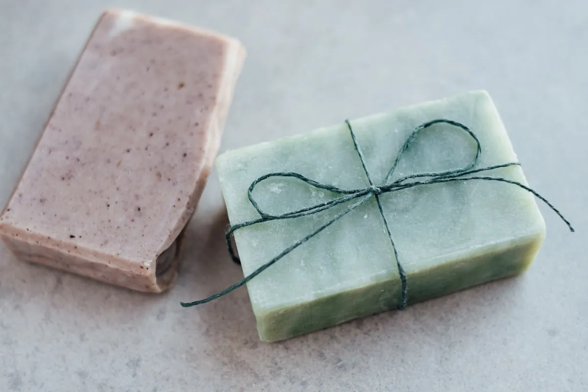 Two handmade soap bars — one green wrapped in dark twine and one pink speckled — on a light grey stone surface