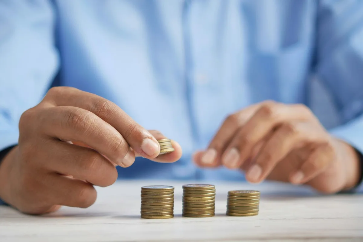 Close-up of two hands building three stacks of coins of visibly different heights on a pale wooden table