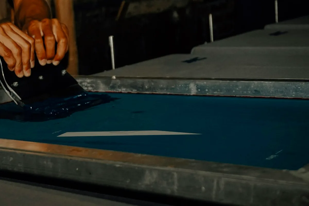 A printer's hands drawing a squeegee through a pool of dark blue ink on a screen frame, with a pale wedge of the stencil showing through the mesh where the design will print