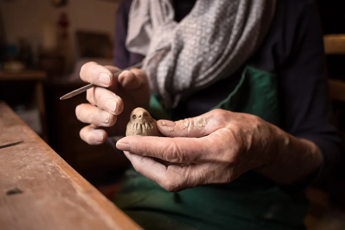 A potter's weathered hands gently shape a small clay bird figurine on a wooden workbench, holding a fine sculpting tool in the right hand, with a green apron and softly lit studio in the background