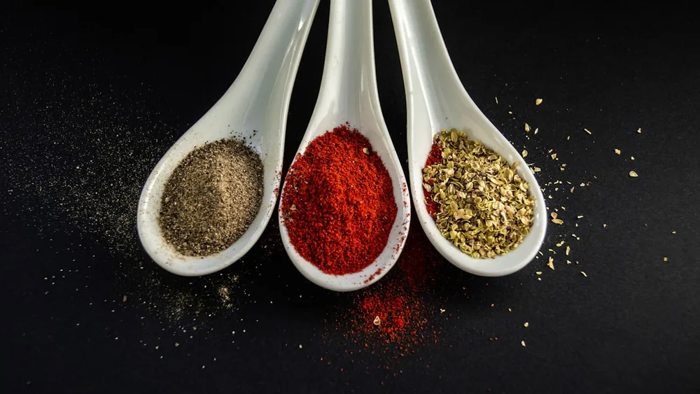 Three white spoons holding ground black pepper, red paprika, and dried oregano against a dark background