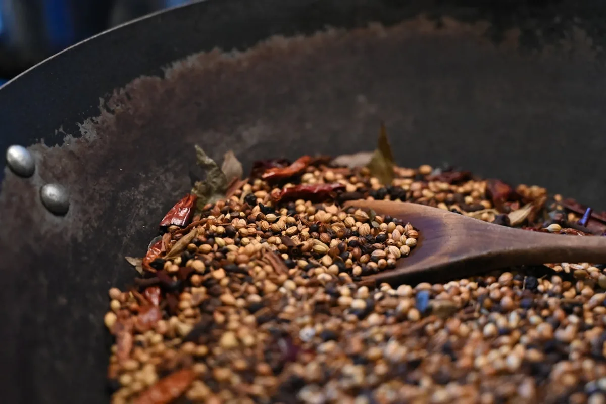 Whole spices — coriander seed, peppercorns, dried chilies, and bay leaves — being mixed with a wooden spoon in a large dark blending pan