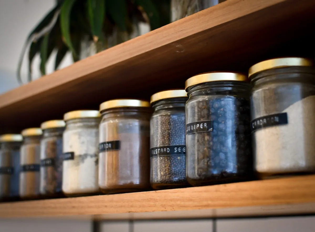 Labeled glass jars of spices with gold lids lined up on a wooden shelf, including juniper, mustard seed, and cardamom