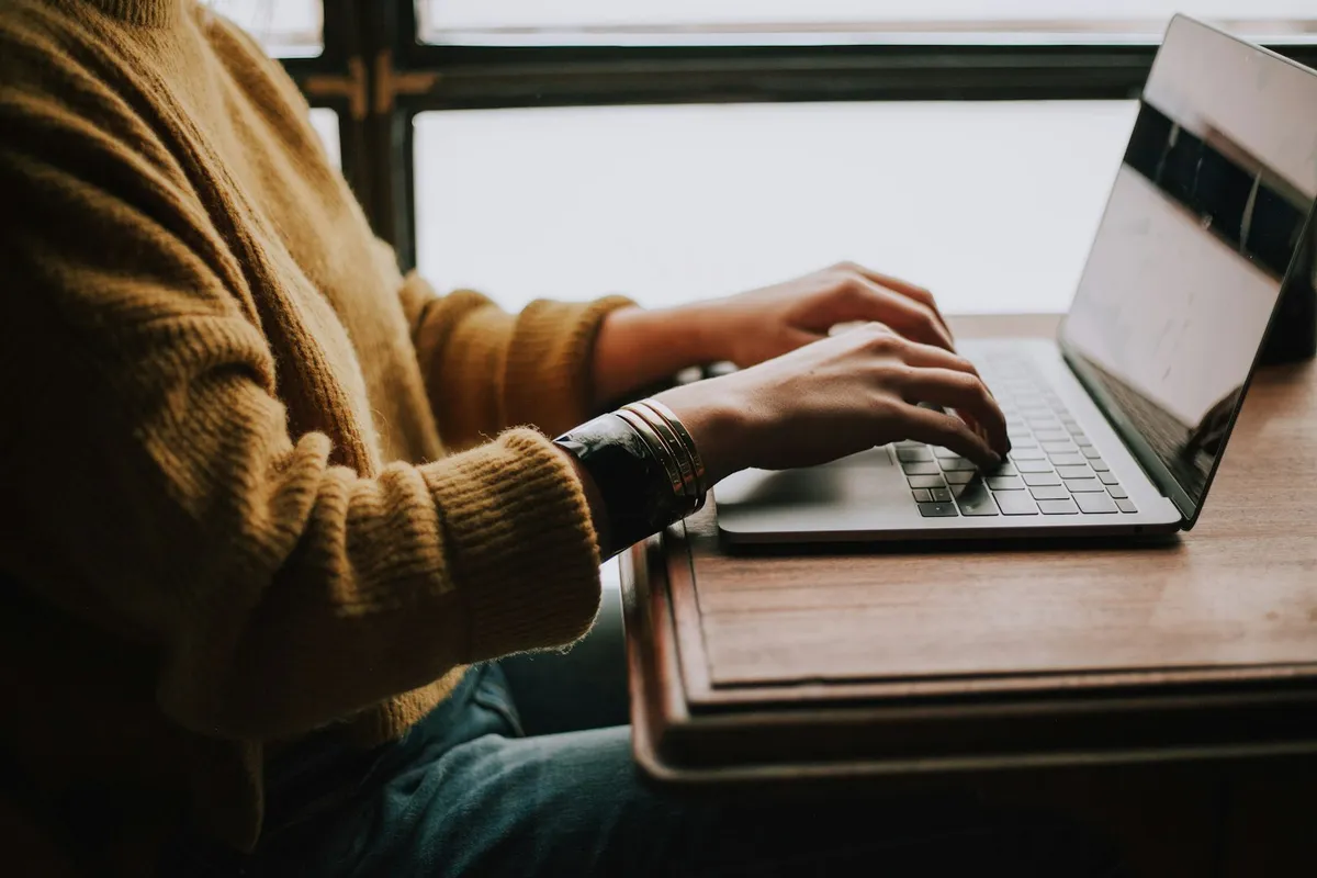 A person in a mustard yellow cable-knit sweater typing on a silver laptop at a wooden desk near a bright window in warm natural light