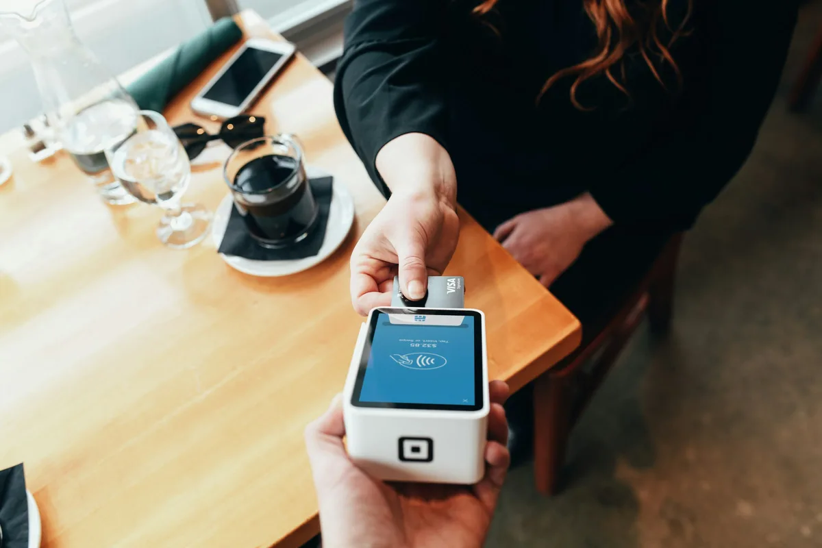 Overhead view of a customer tapping a Visa card against a white countertop card terminal held by a seller, beside a coffee cup and water glasses on a wooden table